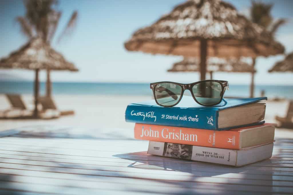 Resort packing list - image shows 3 books and a pair of sunglasses on a table. Behind you see the ocean and some chairs and beach umbrellas.