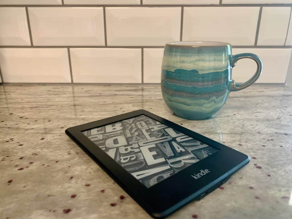 A kindle reader (a travel must-have) and a teal mug on a white and grey countertop with metro tiles backsplash behind.