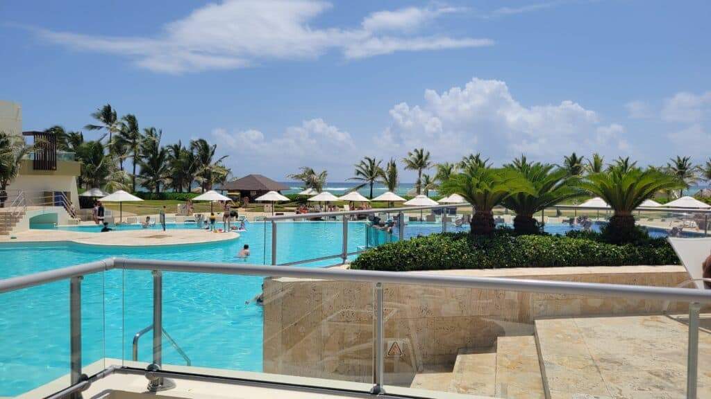 A pool with several white umbrellas, some palm trees and a blue ocean in the background at this Dominican Republic all-inclusive family resort