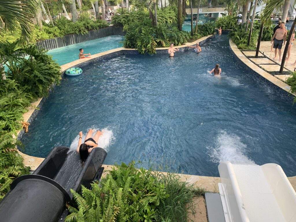 Two water slides in a blue pool, several people in and around the pool, and also some greeneries at this Punta Cana all-inclusive family resort