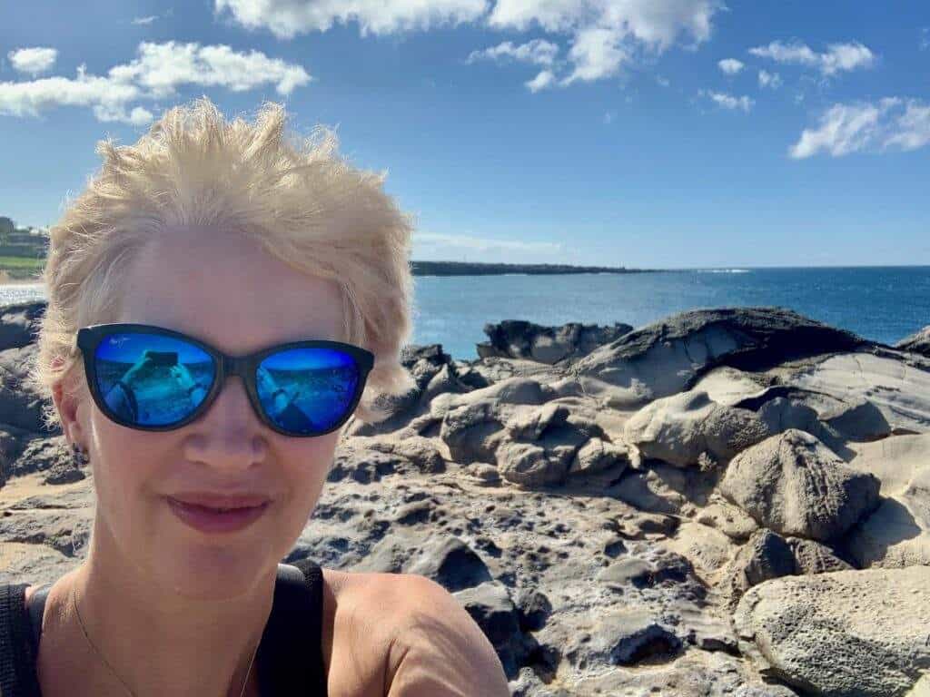 A woman with blond hair wearing sunglasses taking a selfie in front of the ocean in Maui