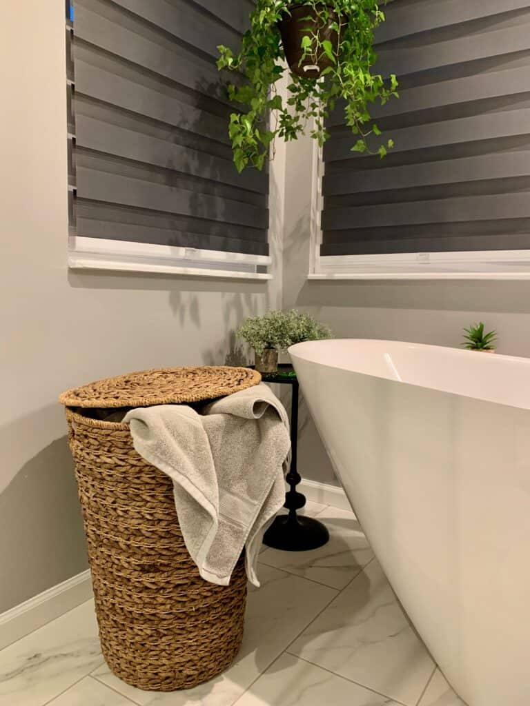 A laundry basket next to a white bathtub with plants surrounding it