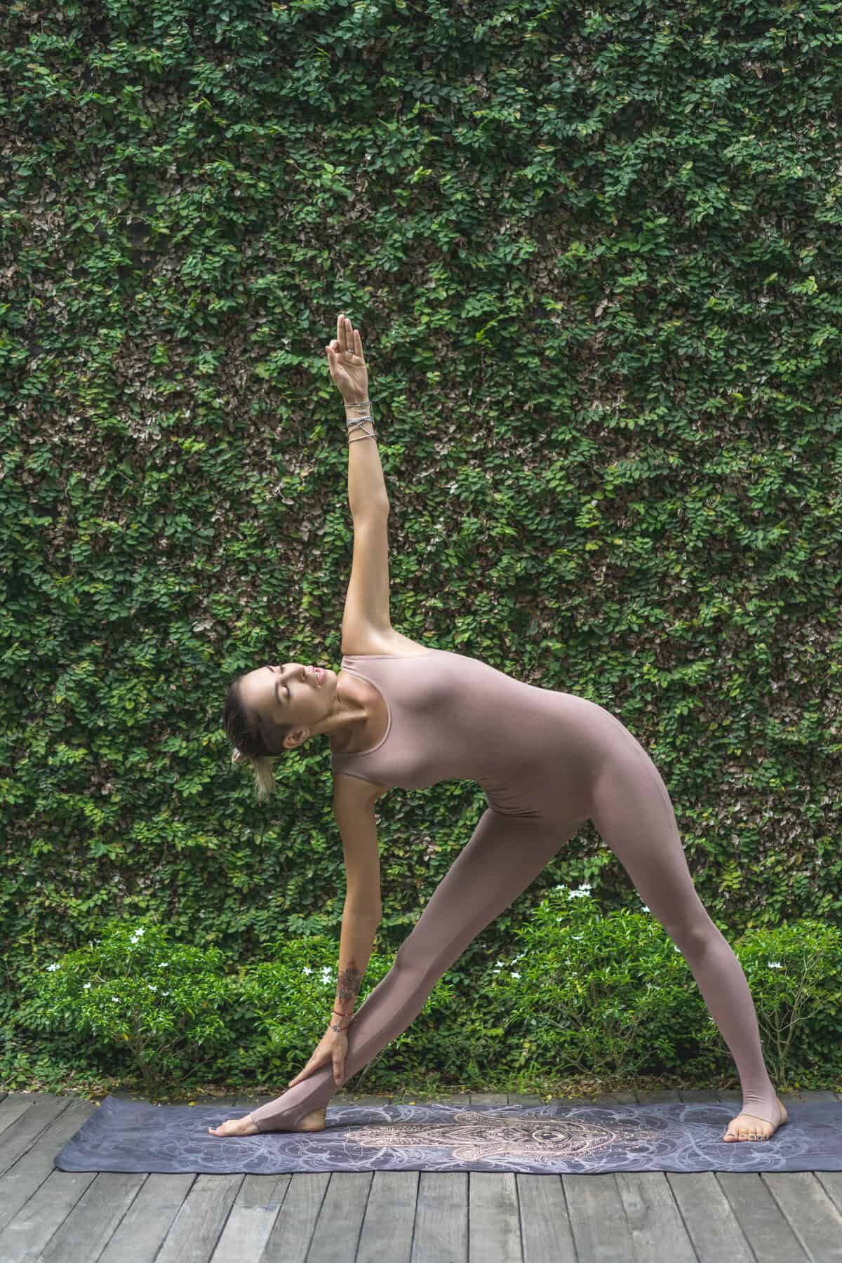 side view of beautiful young woman practicing yoga in Revolved Triangle pose (Parivrtta Trikonasana) in front of wall covered with green leaves to gain one of the 7 benefits of yoga around improved digestion