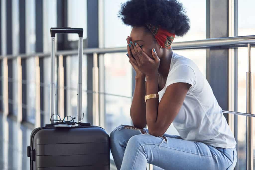 Sad and depressed young african american female passanger in casual clothes is in airport with baggage during air travel disruptions