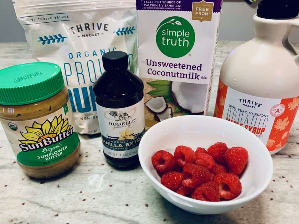 An image of many ingredients to make a healthy quinoa breakfast bowl: sunflower butter, vanilla extract, quinoa, raspberries, maple syrup, and coconut milk.