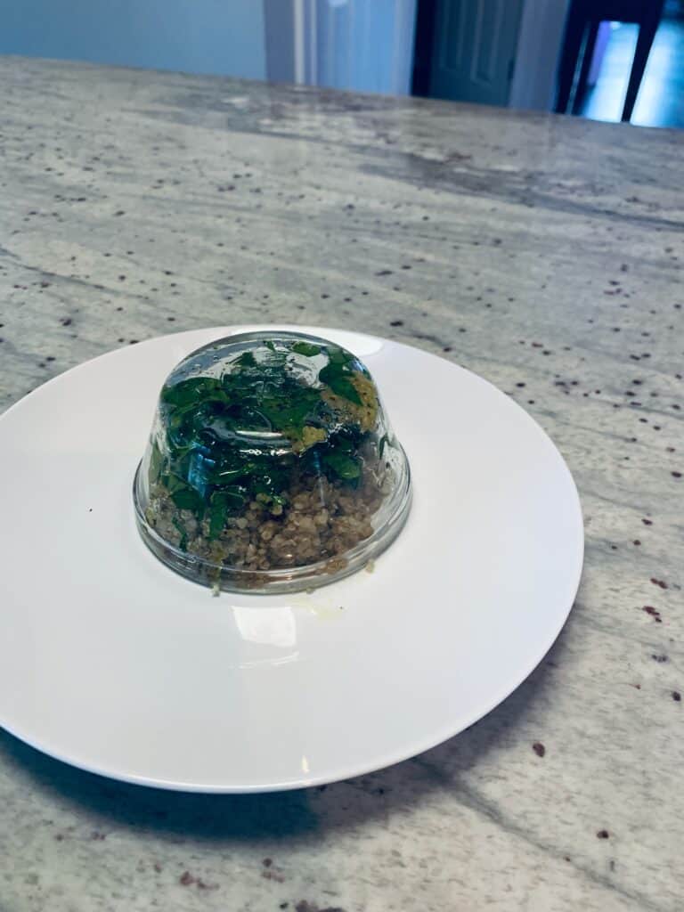 A glass bowl with quinoa and herbs in it sitting upside-down on a white plate.