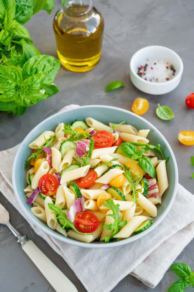 Healthy vegan pasta salad with tomatoes, cucumbers, red onions and arugula in a bowl on a dark gray background. Perfect use of summer vegetables and fruits.