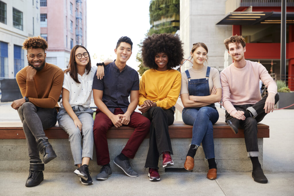 Six young adult friends sitting in a row on a bench in a city street smiling to camera, full length
