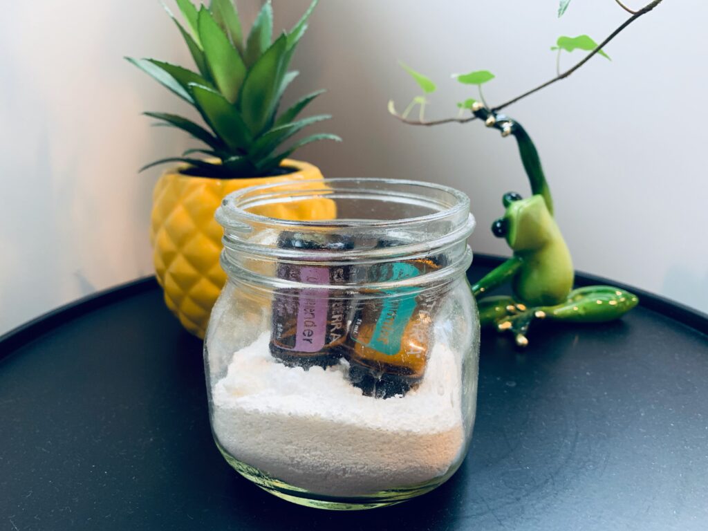 A glass jar of Epsom salt with 2 upside down bottles of essential oils with a frog and a plant behind