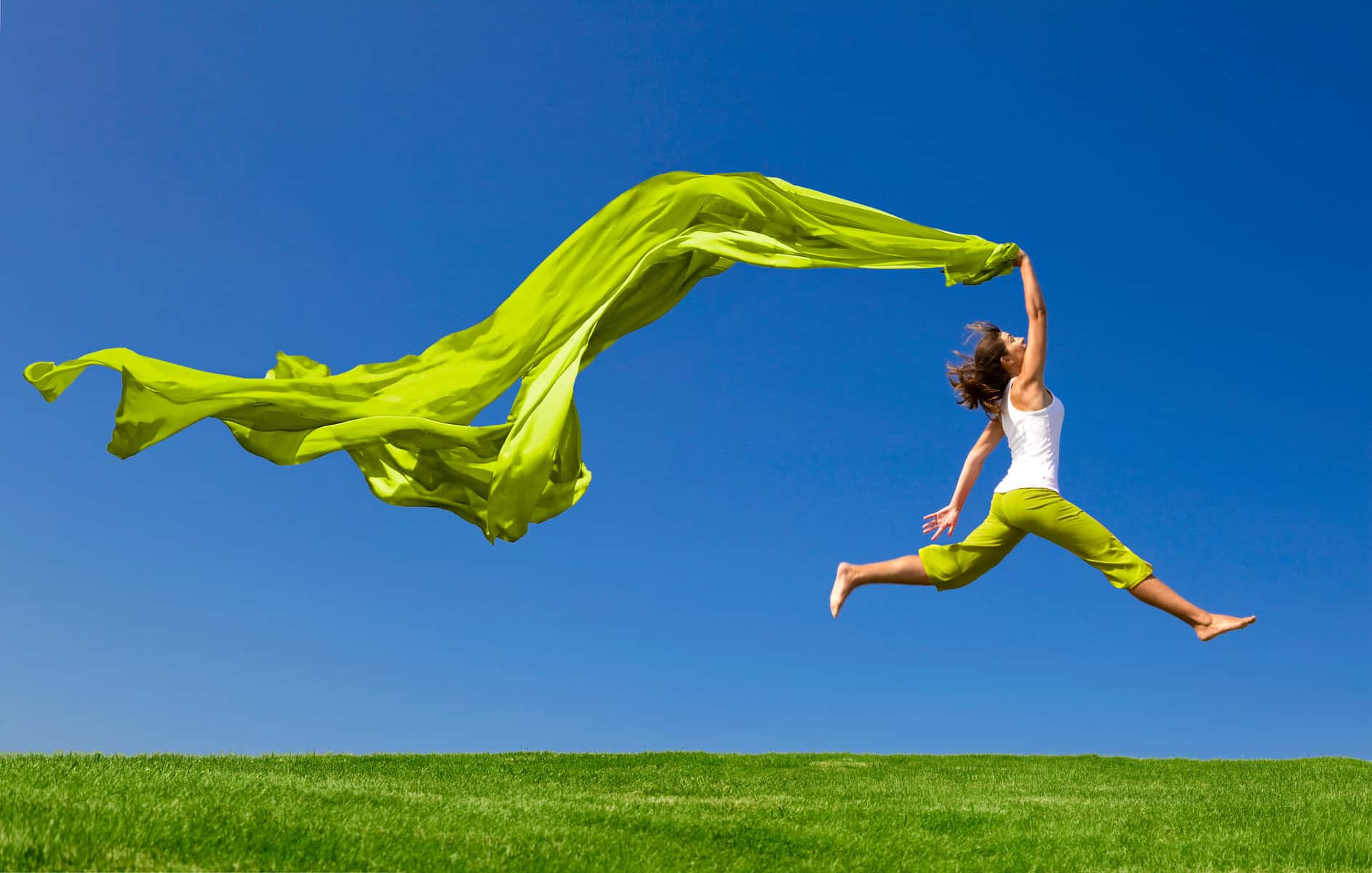 A woman jumping in a grass field holding a long bright green scarf