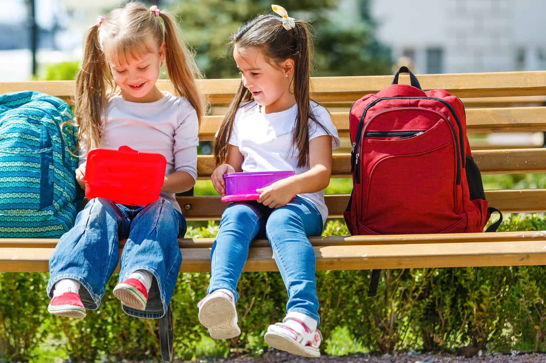 Two young girls sitting on a park bench with their lunch boxes.