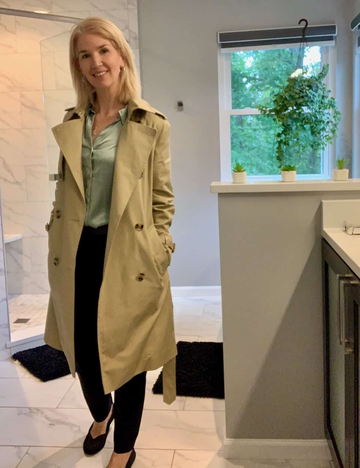 Woman wearing black trousers and black flats, a satin sage shirt and a tan trench coat. Great professional fall capsule wardrobe pieces.