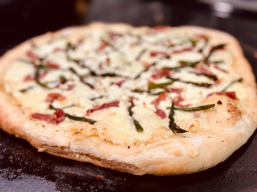 A homemade pizza on a pizza stone with asparagus and prosciutto on it