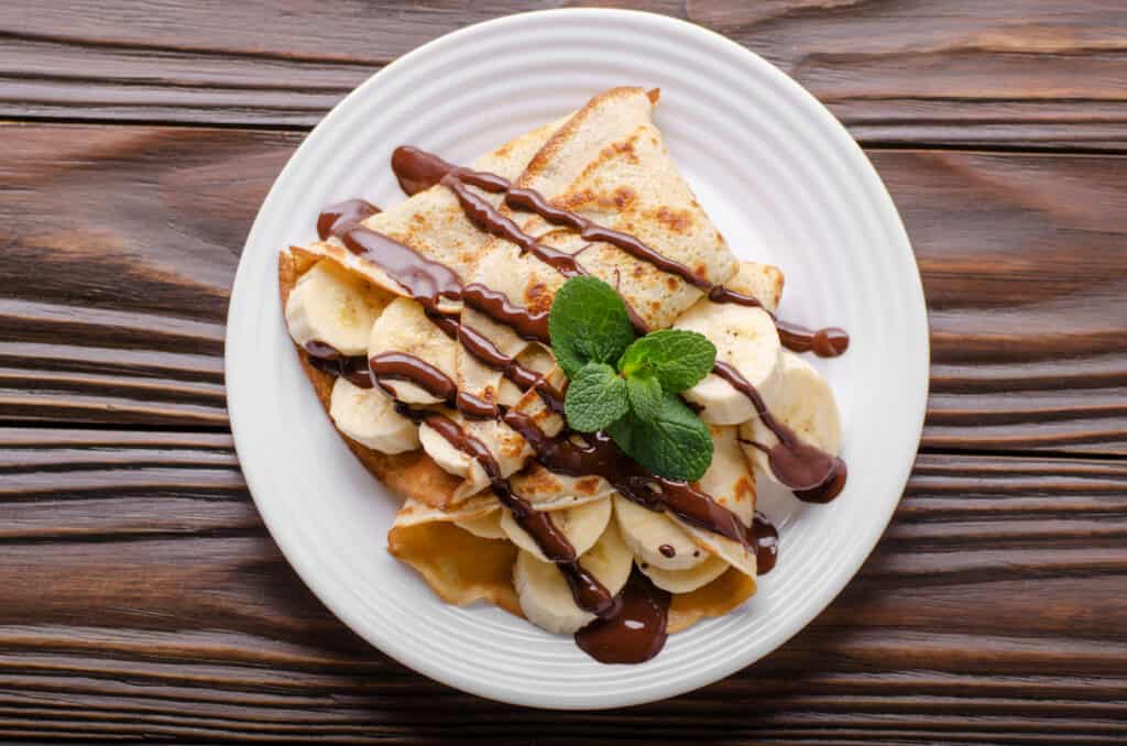 Flat lay French crepes with chocolate sauce and banana in ceramic dish on wooden kitchen table perfect for a brunch buffet